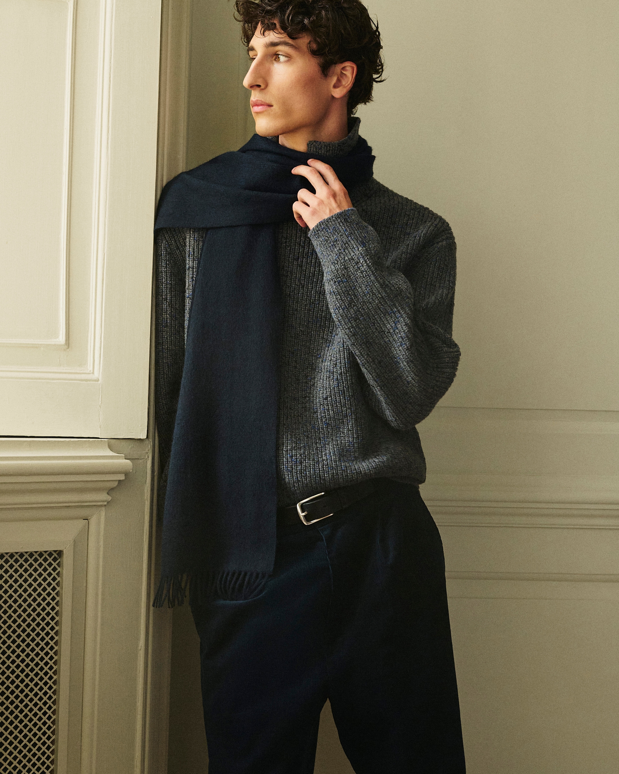 A person in a gray sweater and dark scarf gazes sideways, standing against a light-colored wall with ornate molding.