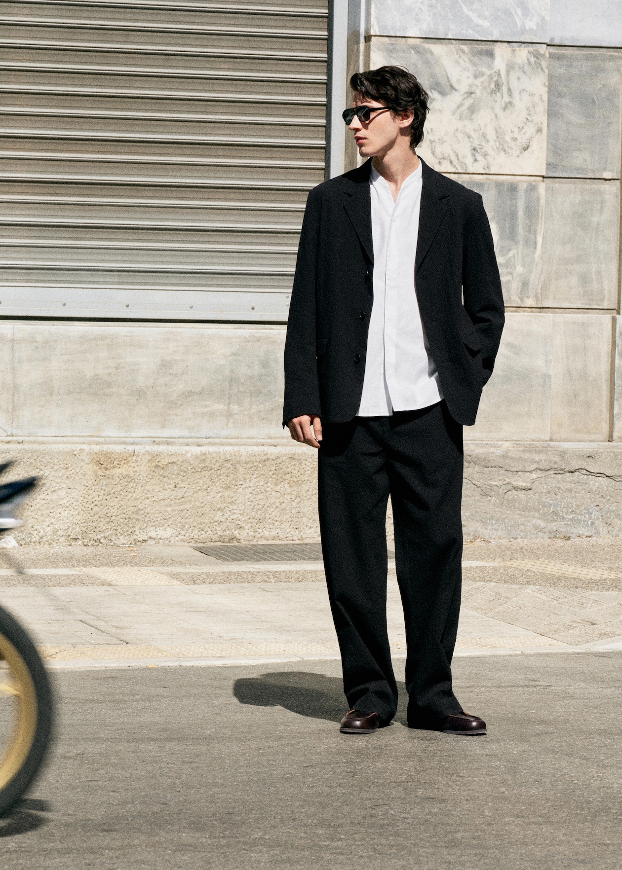 Man in black suit and white shirt, wearing sunglasses, stands on a street with a blurred bicycle passing by, near a stone buildingg.