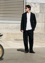 Placeholder for: Man in black suit and white shirt, wearing sunglasses, stands on a street with a blurred bicycle passing by, near a stone buildingg.