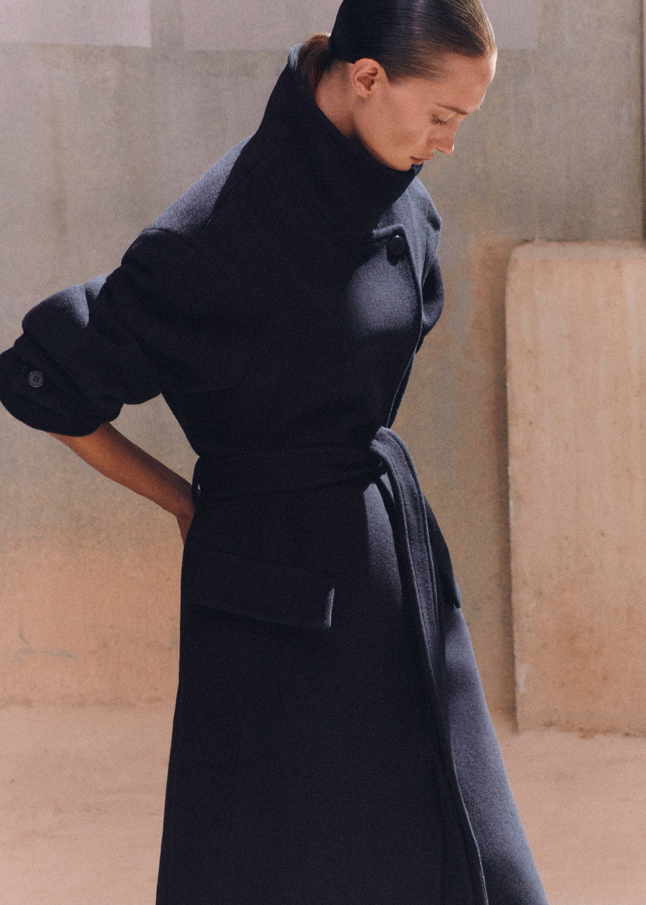 Person wearing a dark, long coat with a high collar and rolled sleeves, looking down in a minimalistic setting.
