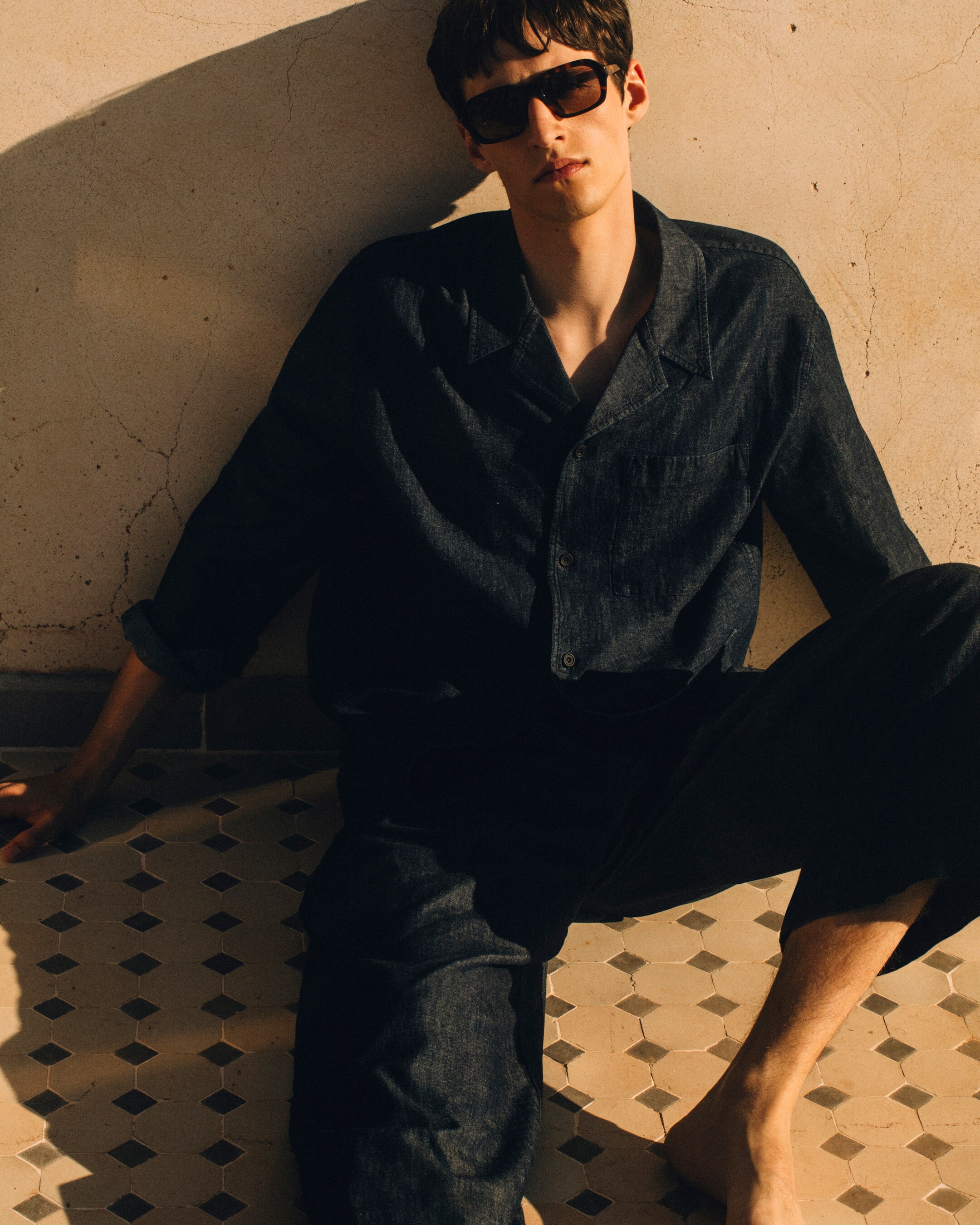 Man in textured co-ord with sunglasses on sitting on the floor against a wall