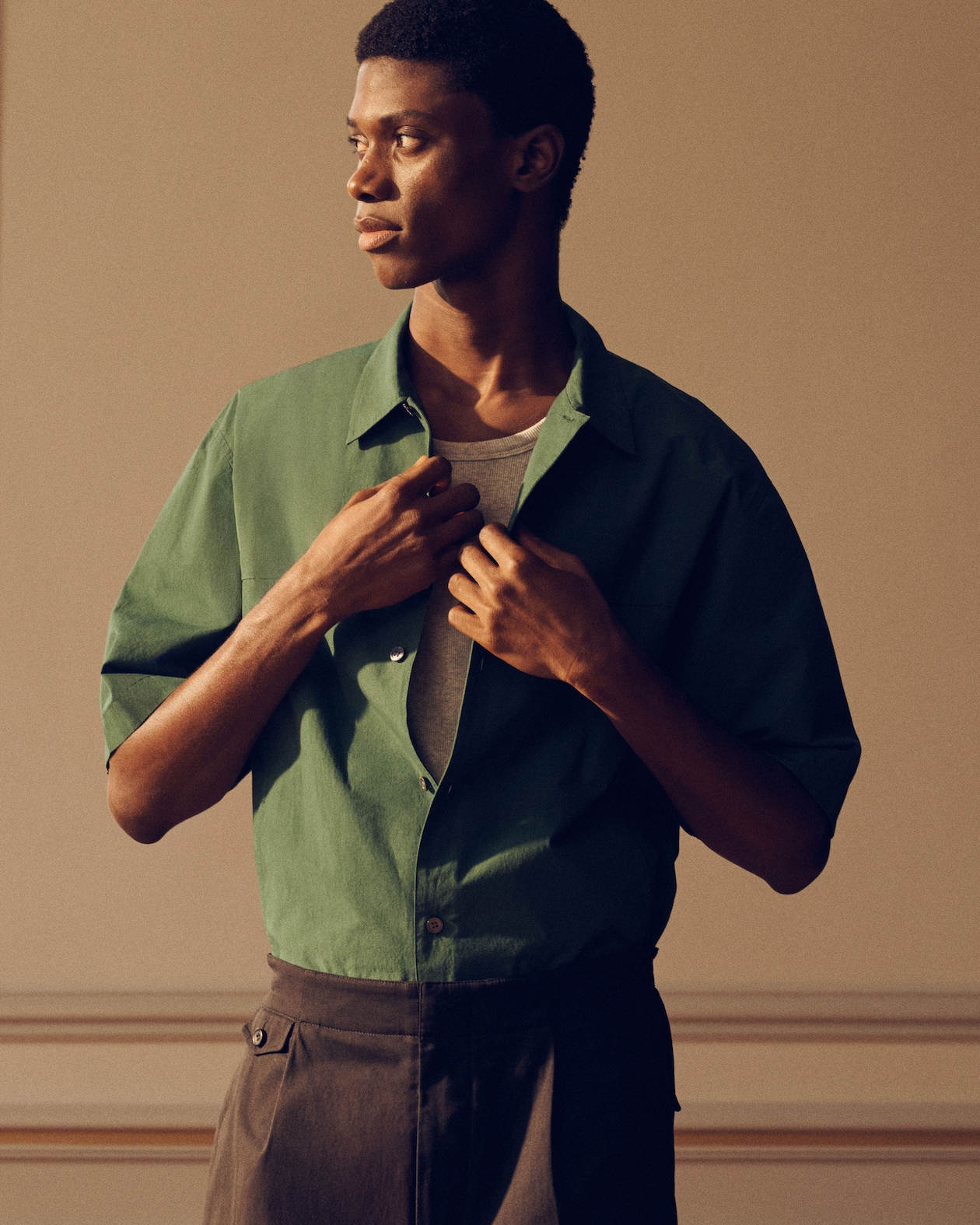 A person in profile wearing a green shirt over a gray top, adjusting the collar. The background is softly lit with warm tones.