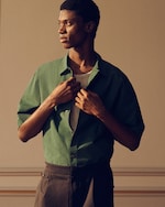 Placeholder for: A person in profile wearing a green shirt over a gray top, adjusting the collar. The background is softly lit with warm tones.