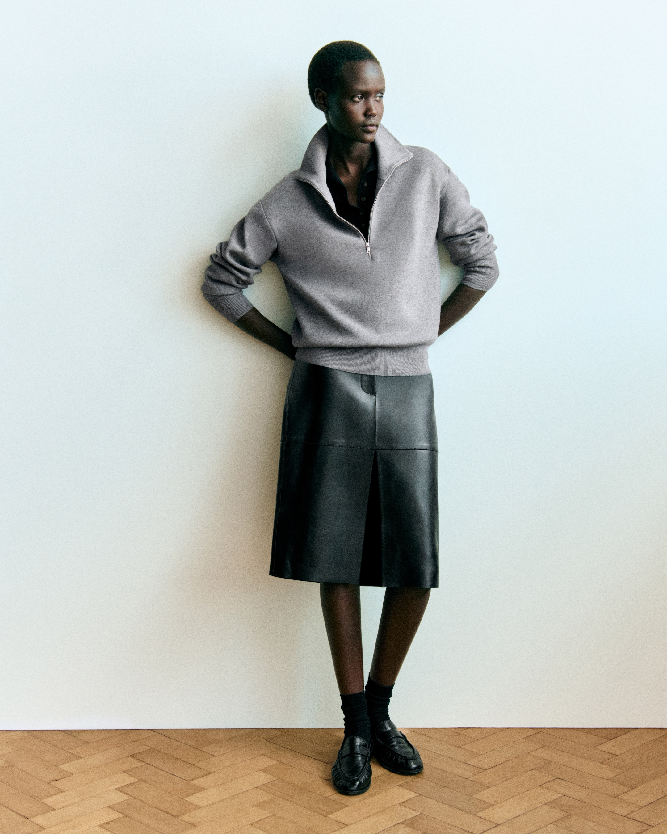 A person in a gray sweater and black leather skirt, standing against a light blue wall with hands behind their back.