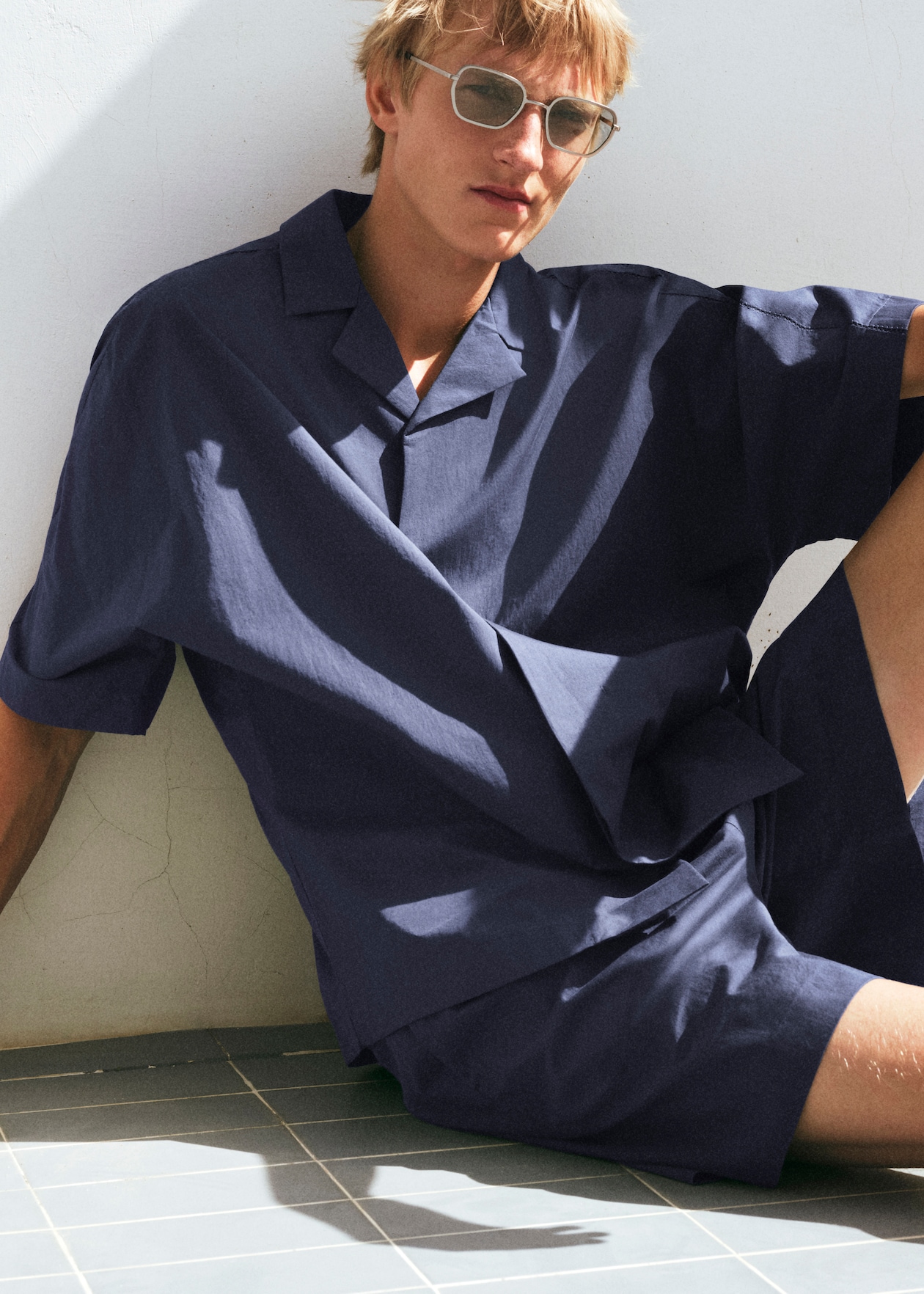 A person wearing sunglasses and a loose navy shirt sits casually against a white wall, with sunlight casting shadows on the tiled floor.