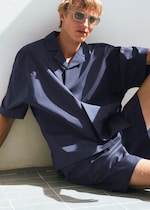 Placeholder for: A person wearing sunglasses and a loose navy shirt sits casually against a white wall, with sunlight casting shadows on the tiled floor.