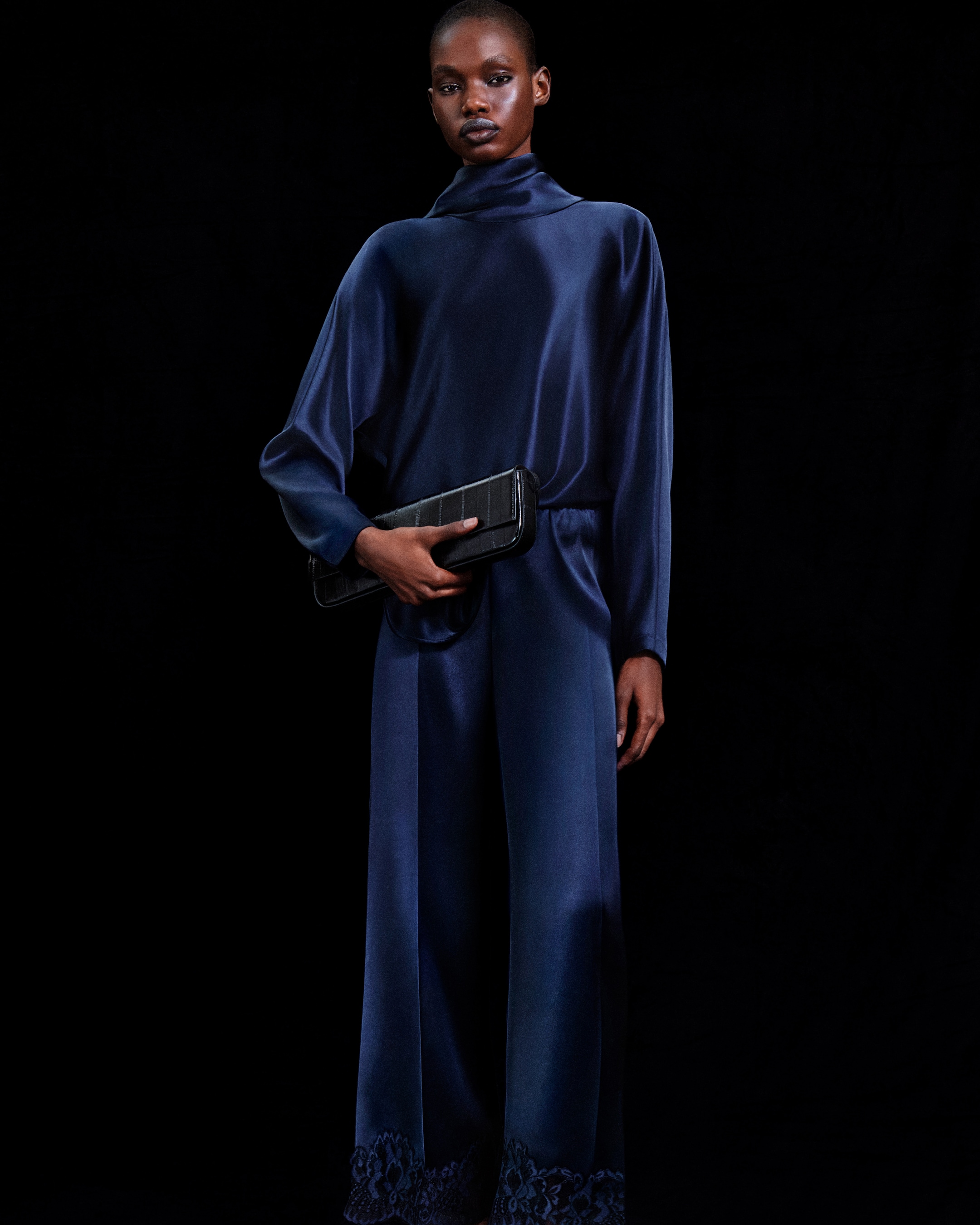 A person in a dark blue satin outfit holding a black clutch, standing against a black background.