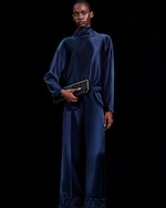 Placeholder for: A person in a dark blue satin outfit holding a black clutch, standing against a black background.