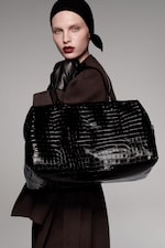 Placeholder for: A person wearing a black headscarf, red lipstick, and brown coat holds a large black crocodile pattern handbag and black gloves.