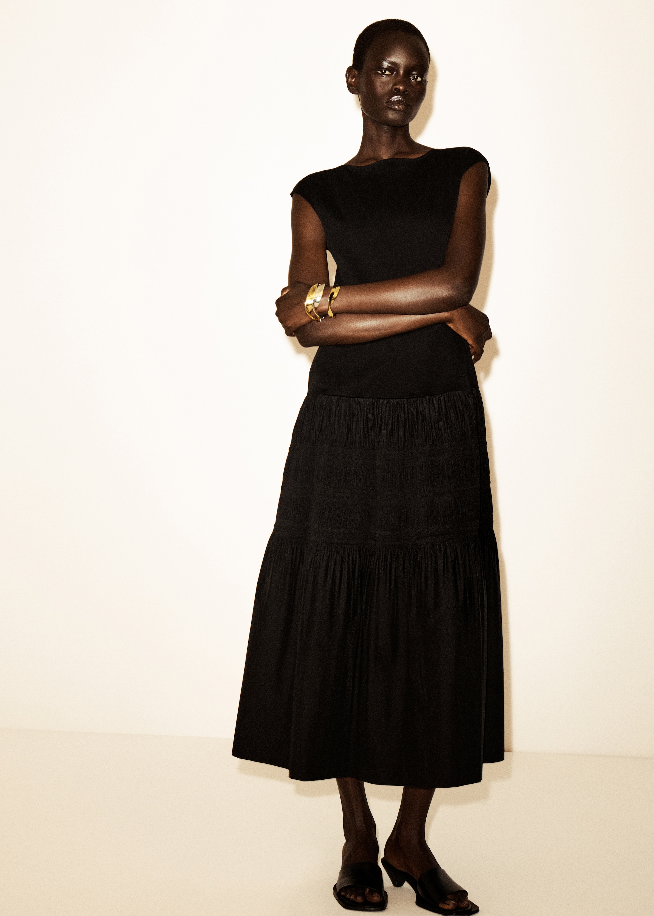 Person in a black dress stands with arms crossed, wearing gold bangles, black open-toe shoes, against a plain background.
