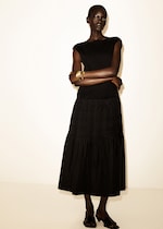 Placeholder for: Person in a black dress stands with arms crossed, wearing gold bangles, black open-toe shoes, against a plain background.