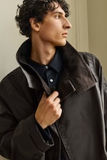 Placeholder for: A person with curly hair wears a dark coat and shirt, holding the collar up while looking to the side.