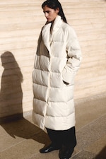 Placeholder for: Person wearing a long, oversized white puffer coat, standing in front of a textured beige wall casting a shadow.