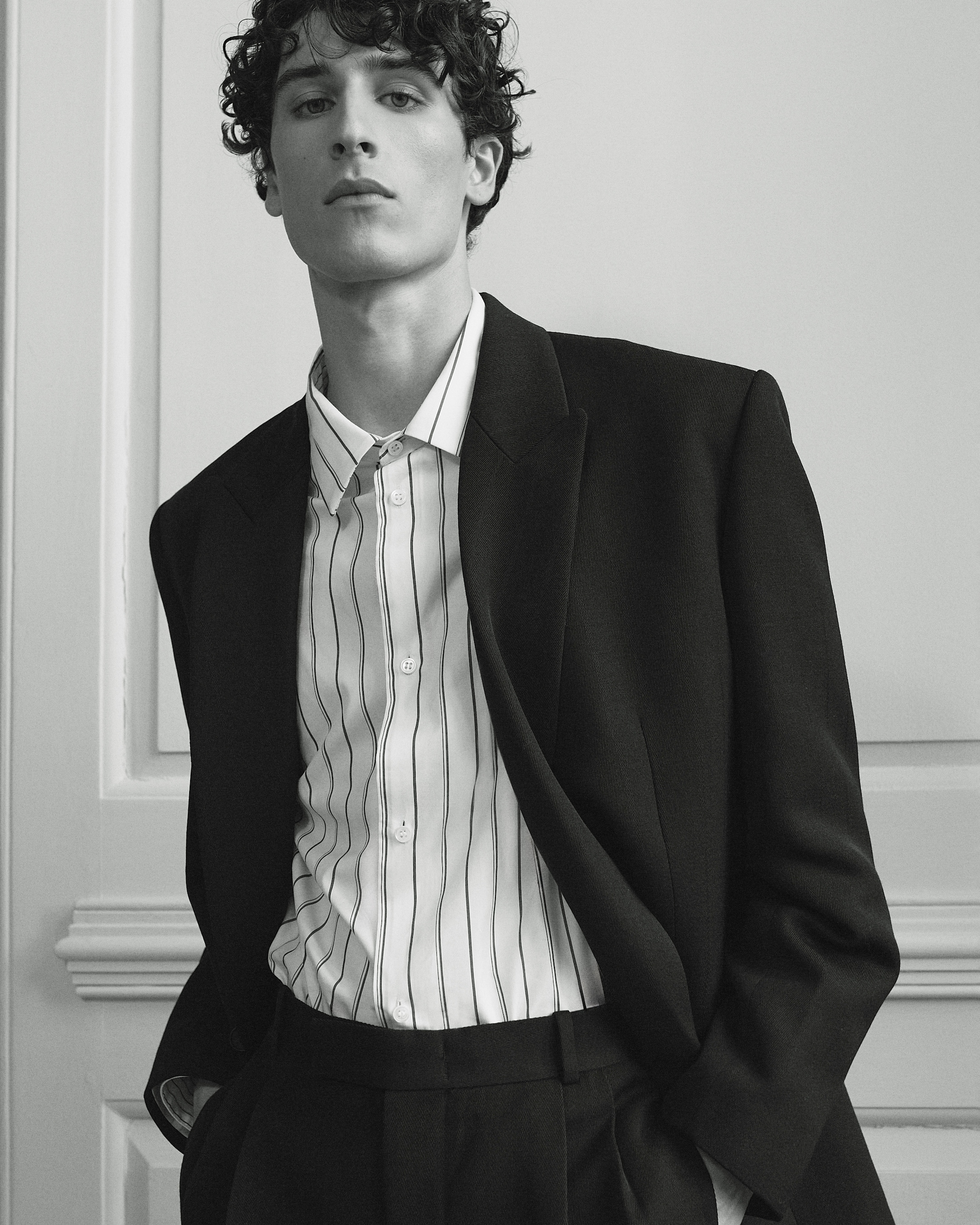 A person with curly hair wearing a dark suit and striped shirt, standing against a light-colored wall, with hands in pant pockets.