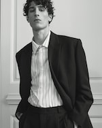 Placeholder for: A person with curly hair wearing a dark suit and striped shirt, standing against a light-colored wall, with hands in pant pockets.