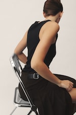 Placeholder for: Person with short hair in a black sleeveless top and dark skirt sits sideways on a metal chair against a plain background.