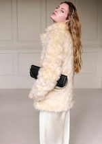 Placeholder for: A woman with long hair wearing a light fur coat holds a black clutch, standing in a softly lit room with paneled walls.