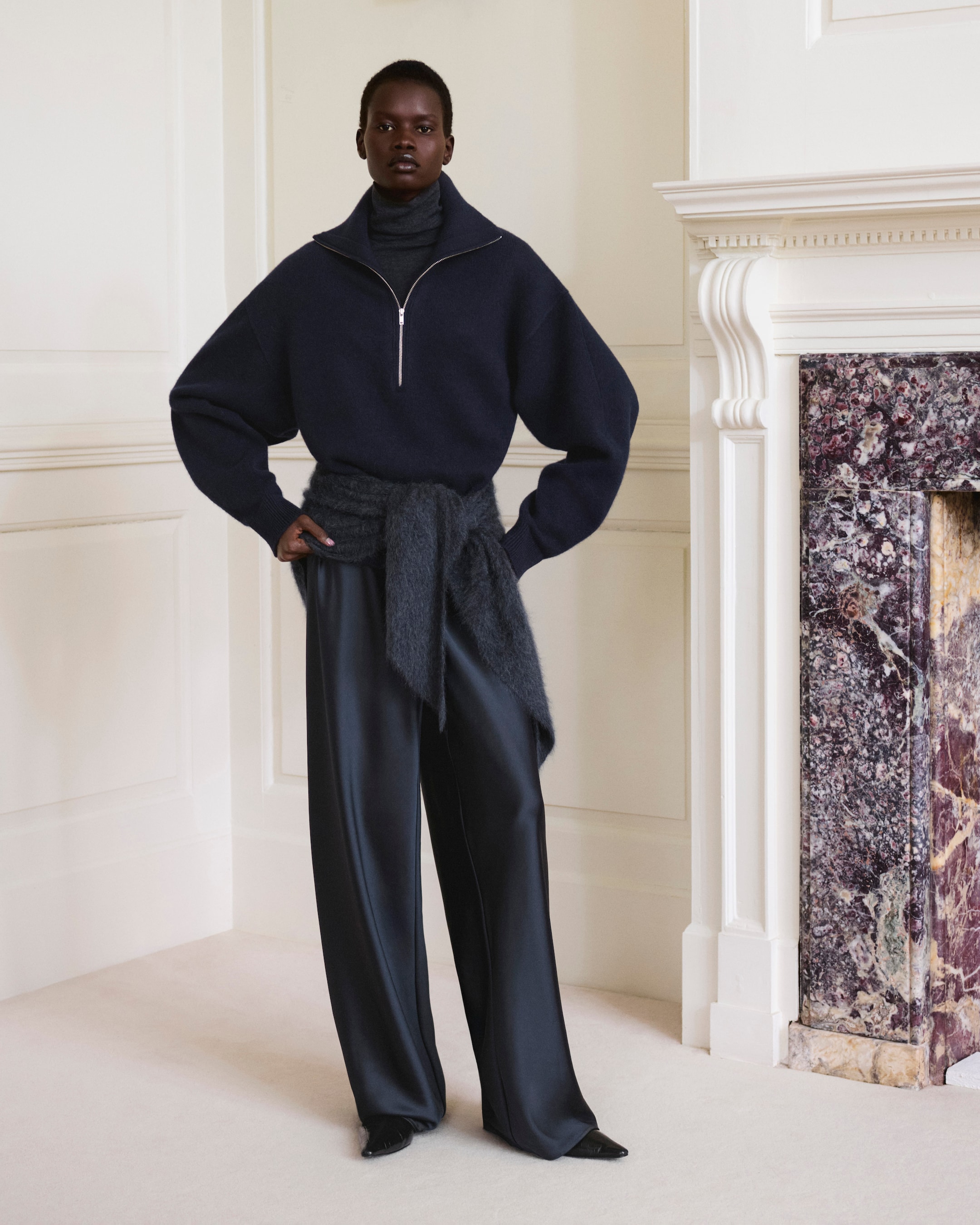 Model wearing a dark sweater, wide trousers, and wrapped scarf, standing in a white room beside a marble fireplace.
