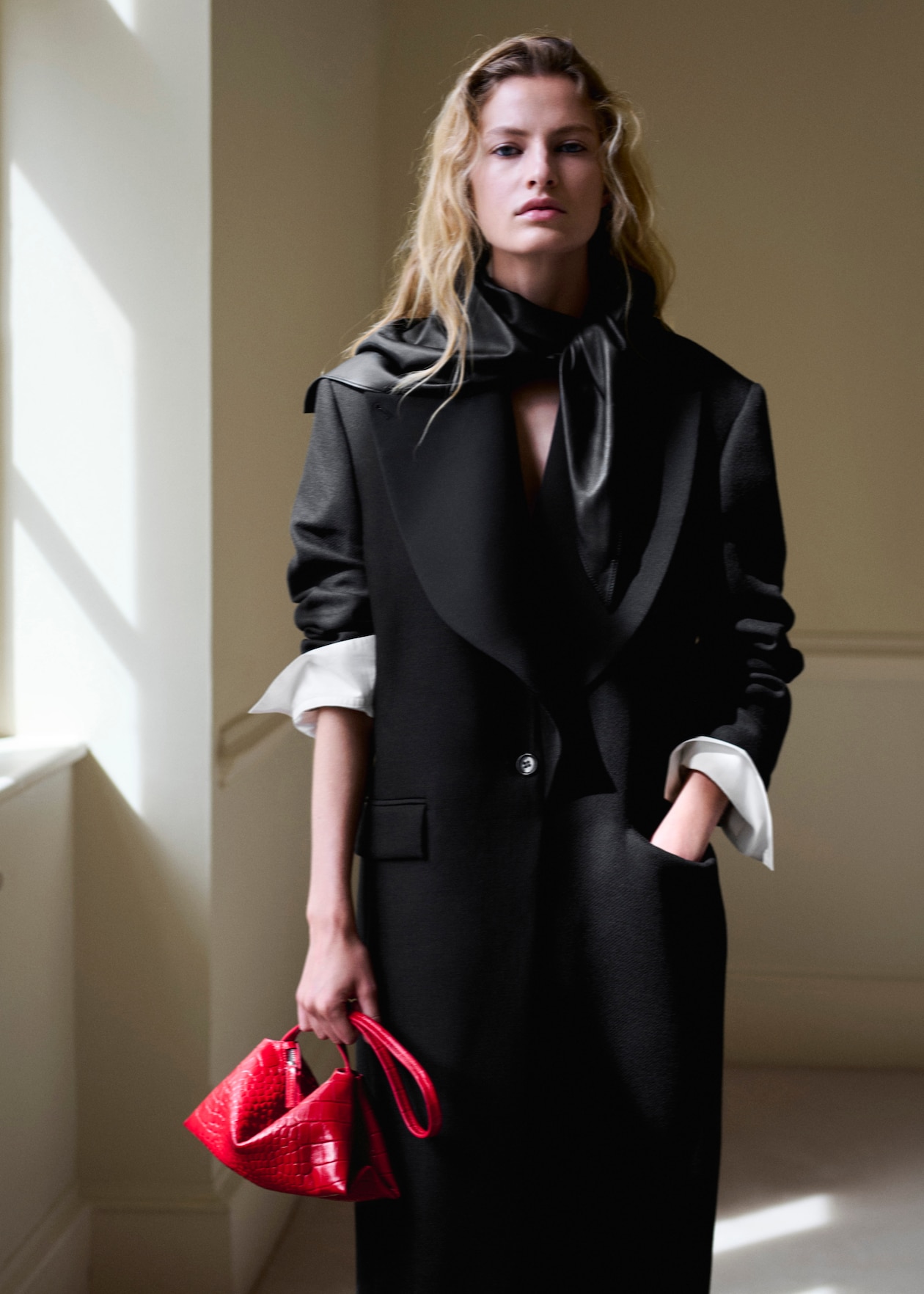 Person with long hair wearing a black coat and holding a small red handbag, standing in a softly lit room.