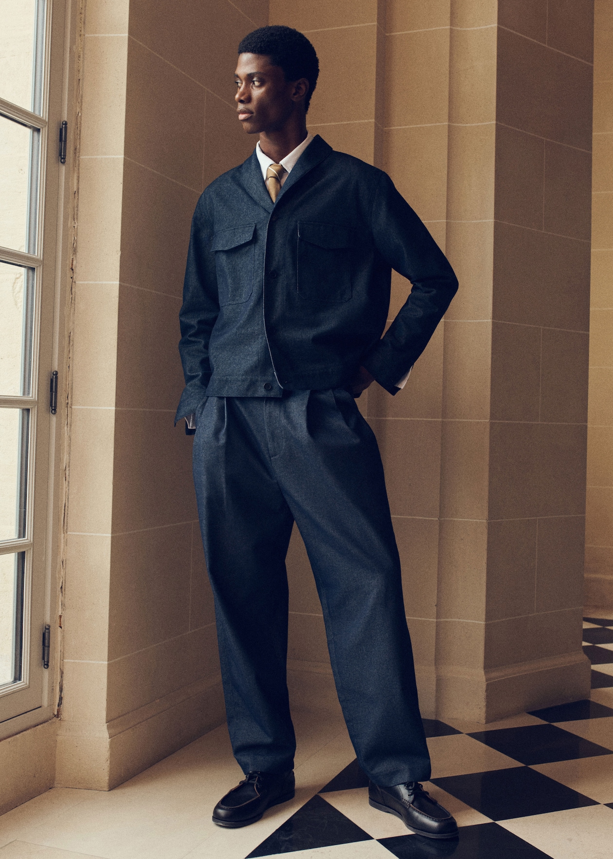 A person in a stylish dark suit with a tie stands by a window, hands in pockets, against a geometric-patterned tiled floor.