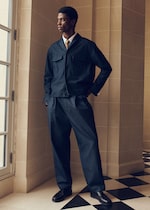 Placeholder for: A person in a stylish dark suit with a tie stands by a window, hands in pockets, against a geometric-patterned tiled floor.