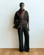 Placeholder for: A person stands against a plain background wearing a dark outfit with a scarf, holding a large bag in one hand, and standing on a wooden floor.