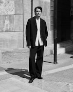 Placeholder for: A man in a white shirt and dark suit stands on a city street, bathed in sunlight, against a textured wall. The image is in black and white.