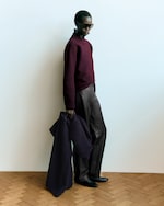 Placeholder for: Person wearing dark sunglasses, a maroon sweater, and gray pants, holding a coat, stands against a plain light background.