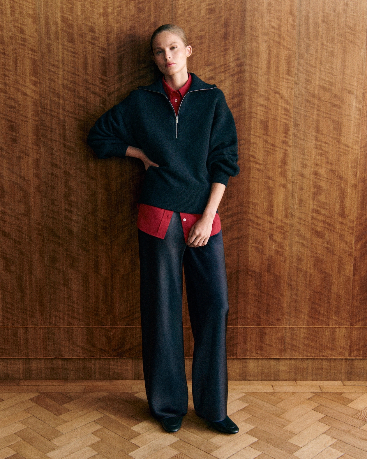 A person leans against a wooden wall, wearing a dark sweater over a red shirt and wide-leg pants, with short hair tied back.