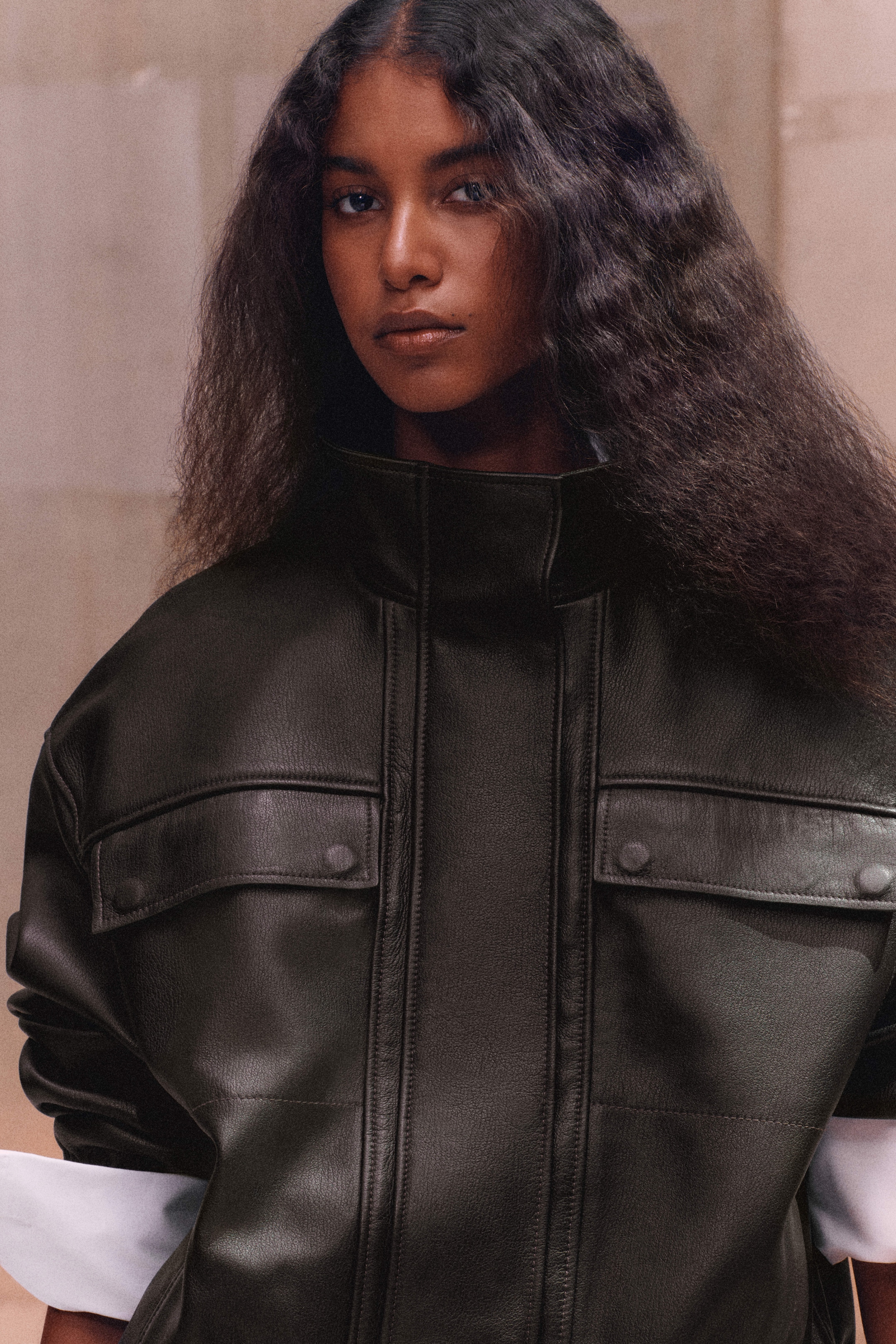 A person with long, textured hair wears a high-collared dark leather jacket, standing against a neutral background.