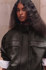 Placeholder for: A person with long, textured hair wears a high-collared dark leather jacket, standing against a neutral background.