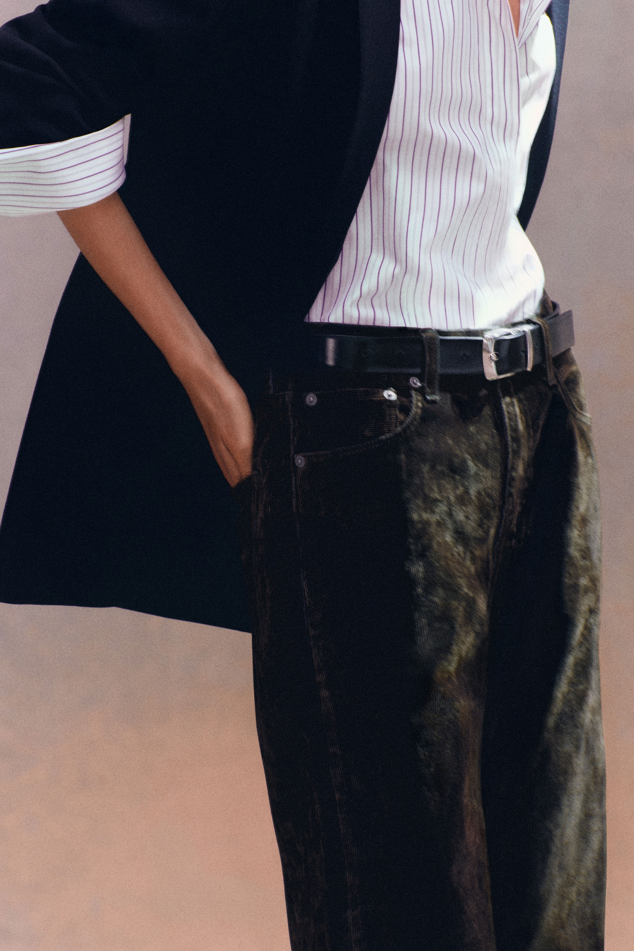 Person wearing a black blazer, striped shirt, and black belt with dark pants, with one hand in the pocket.