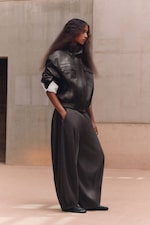 Placeholder for: A person wearing a black leather jacket and wide-leg gray pants, standing in a minimalist indoor setting, looking to the side.
