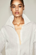 Placeholder for: Person wearing a striped, white collared shirt with an open neckline, looking directly at the camera against a neutral background.