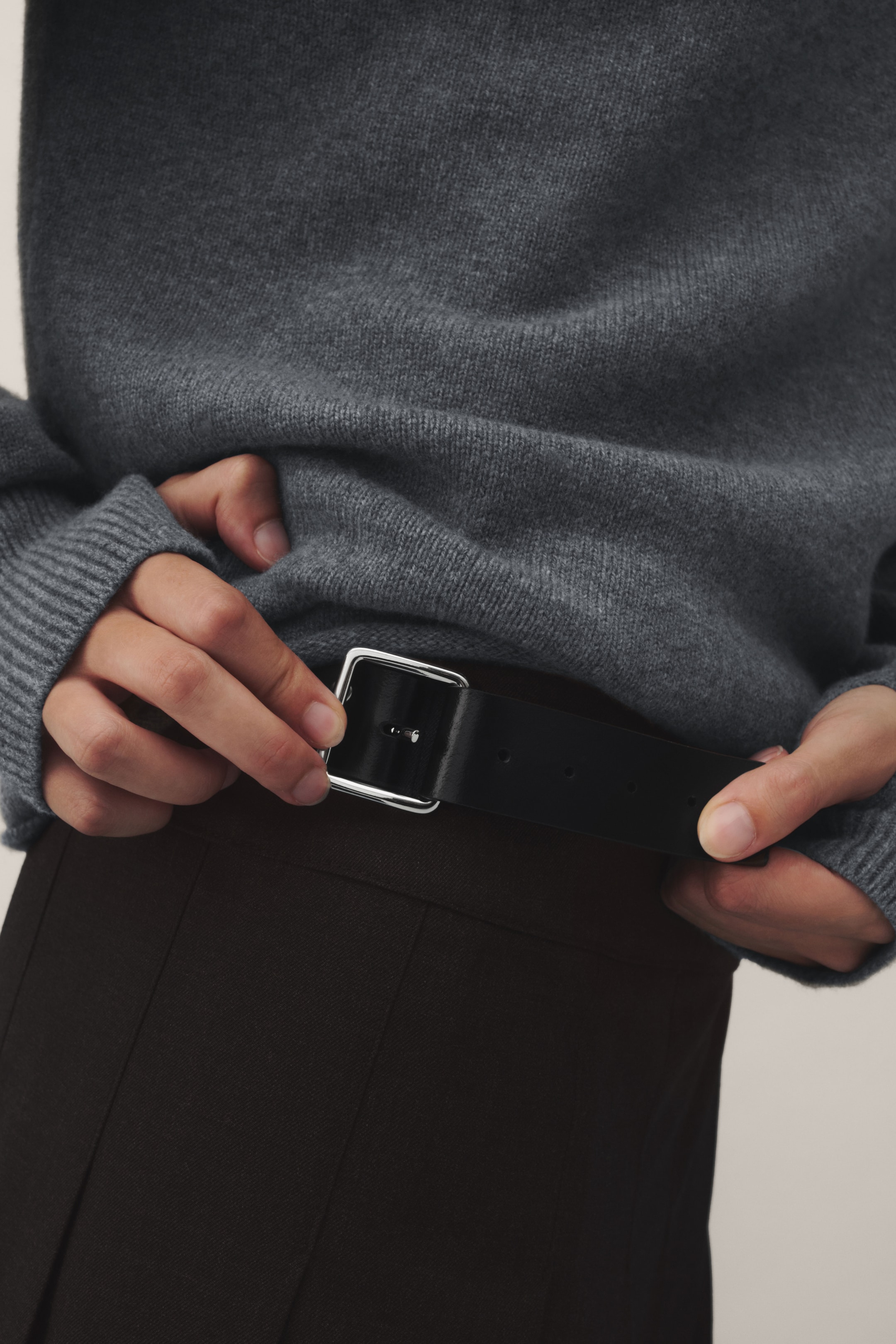 Person wearing a gray sweater and brown pants, adjusting a black belt with a silver buckle.