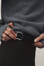 Placeholder for: Person wearing a gray sweater and brown pants, adjusting a black belt with a silver buckle.