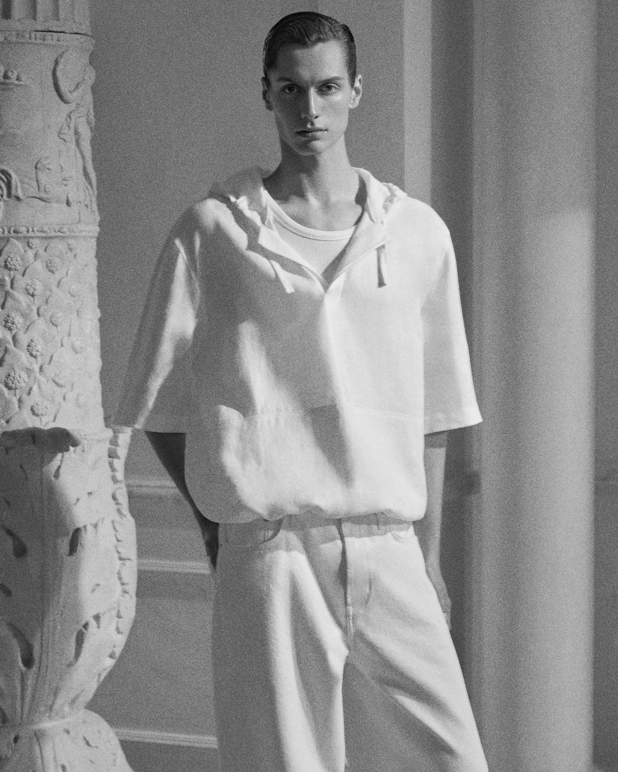 Black and white image of a person in a loose, light outfit, standing beside an ornate, carved pillar indoors.
