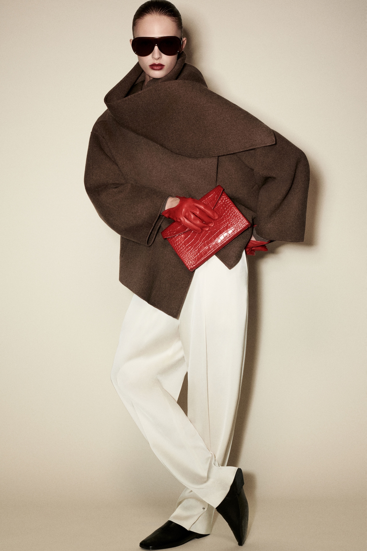 Person wearing oversized brown coat, white trousers, red gloves, holding a red clutch, with dark sunglasses, on a neutral background.