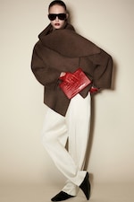 Placeholder for: Person wearing oversized brown coat, white trousers, red gloves, holding a red clutch, with dark sunglasses, on a neutral background.