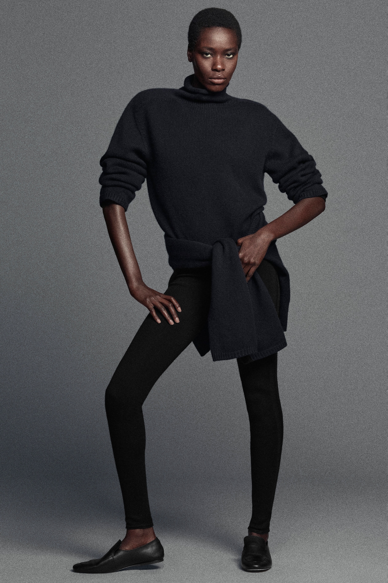 A person wearing a dark turtleneck sweater, black leggings, and black shoes, posing confidently with hands on hips against a plain gray background.