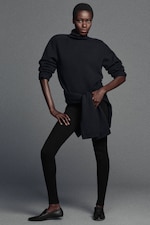 Placeholder for: A person wearing a dark turtleneck sweater, black leggings, and black shoes, posing confidently with hands on hips against a plain gray background.