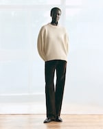 Placeholder for: A person standing indoors wearing a cream sweater and dark pants, with a neutral expression and a softly lit background.