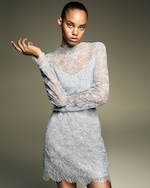 Placeholder for: A person in a light gray lace dress with long sleeves and intricate floral patterns, holding one hand up, against a neutral background.