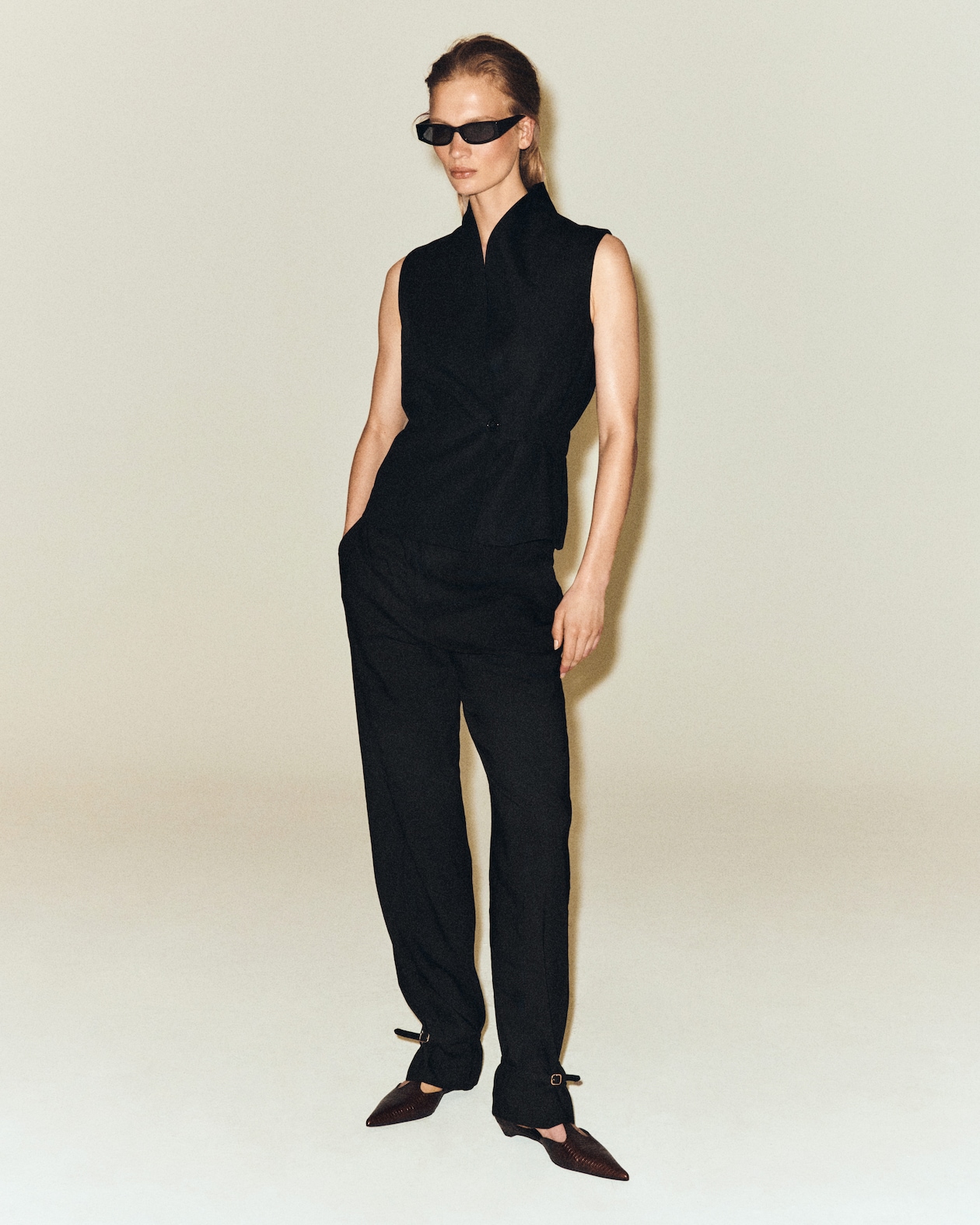 Person wearing a stylish black sleeveless top and pants, sunglasses, and pointed shoes, posing against a plain background.