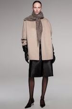 Placeholder for: A person wearing a beige coat, grey scarf, black leather gloves, skirt, hosiery, and heels, standing against a neutral background.