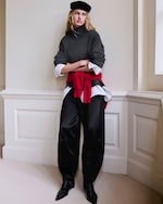 Placeholder for: A woman in a dark turtleneck, white shirt, black pants, and a red sweater tied at the waist, wearing a black beret, stands in a corner with arms crossed.