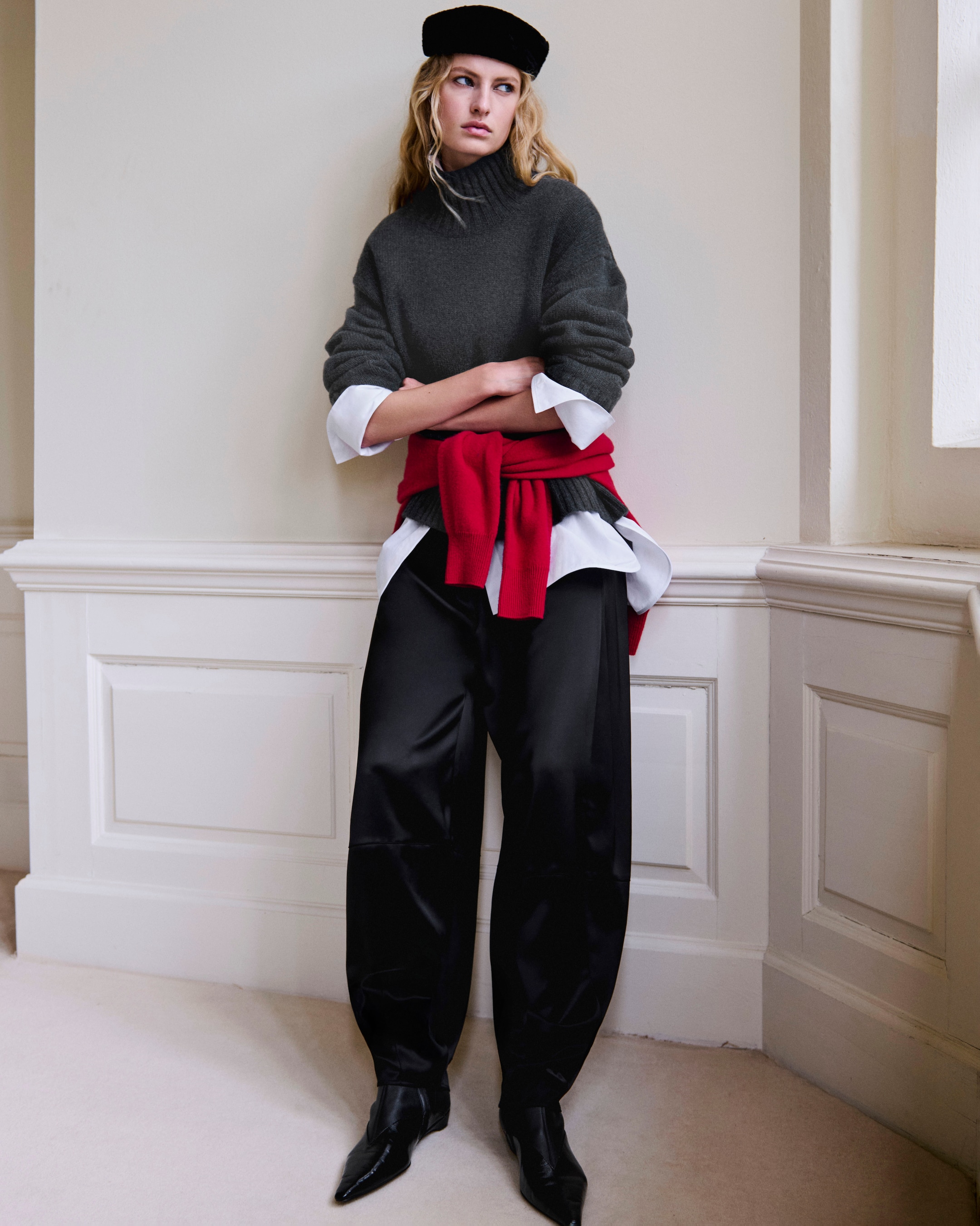 A person leaning against a wall, wearing a gray turtleneck, black pants, red sweater around the waist, and a black beret, with arms crossed.