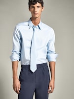 Placeholder for: A person wearing a light blue shirt with rolled-up sleeves, light blue tie, and dark pants stands against a neutral background.