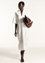 Placeholder for: Woman in a white dress holding a brown clutch, walking confidently against a plain background.