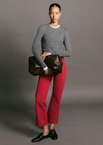 Placeholder for: A person stands holding a large brown clutch, wearing a gray long-sleeve shirt, red pants, and black shoes, against a plain background.